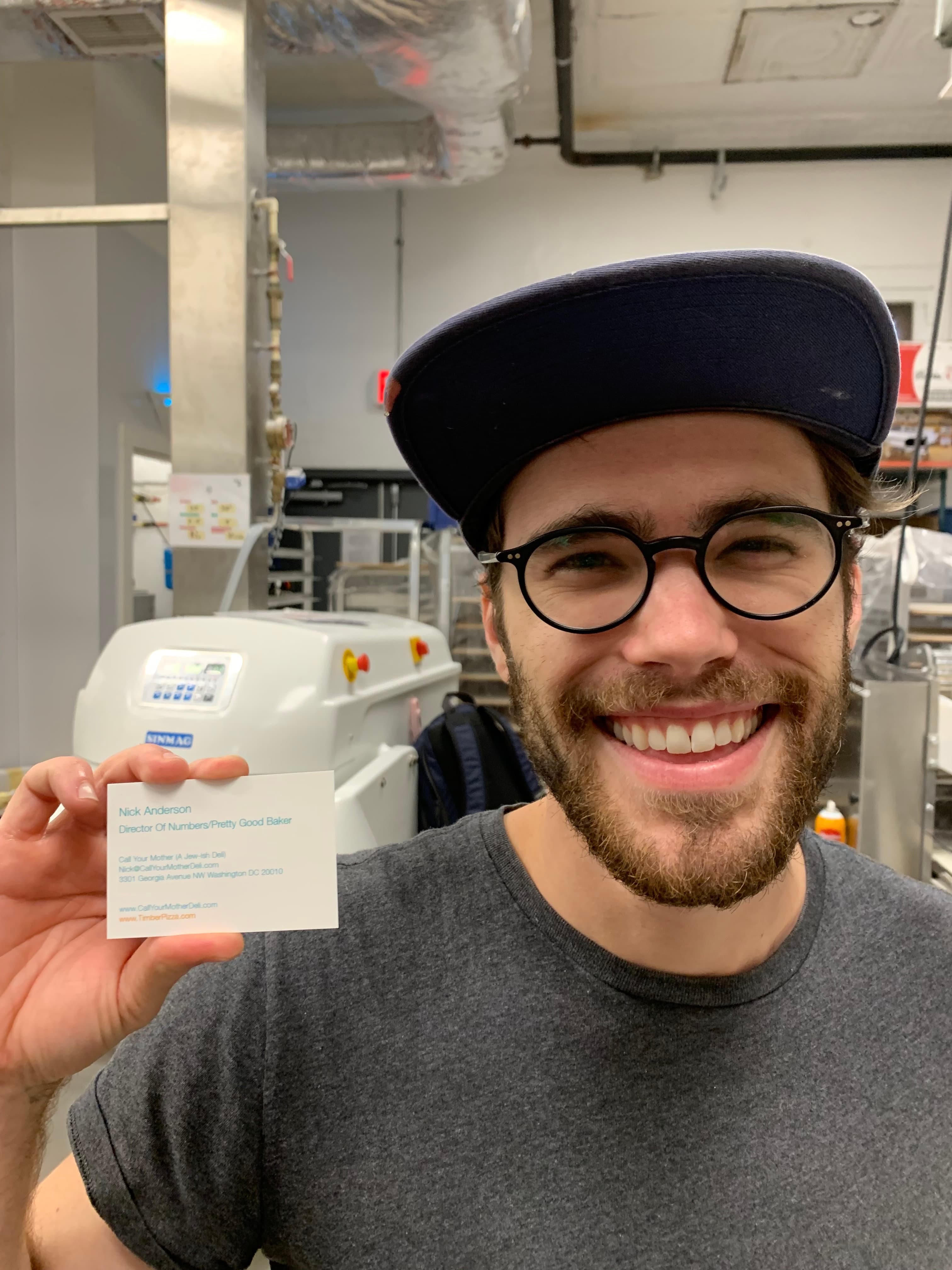 Nick in a bakery kitchen holding his Call Your Mother Deli business card titled 'Director of Numbers / Pretty Good Baker'