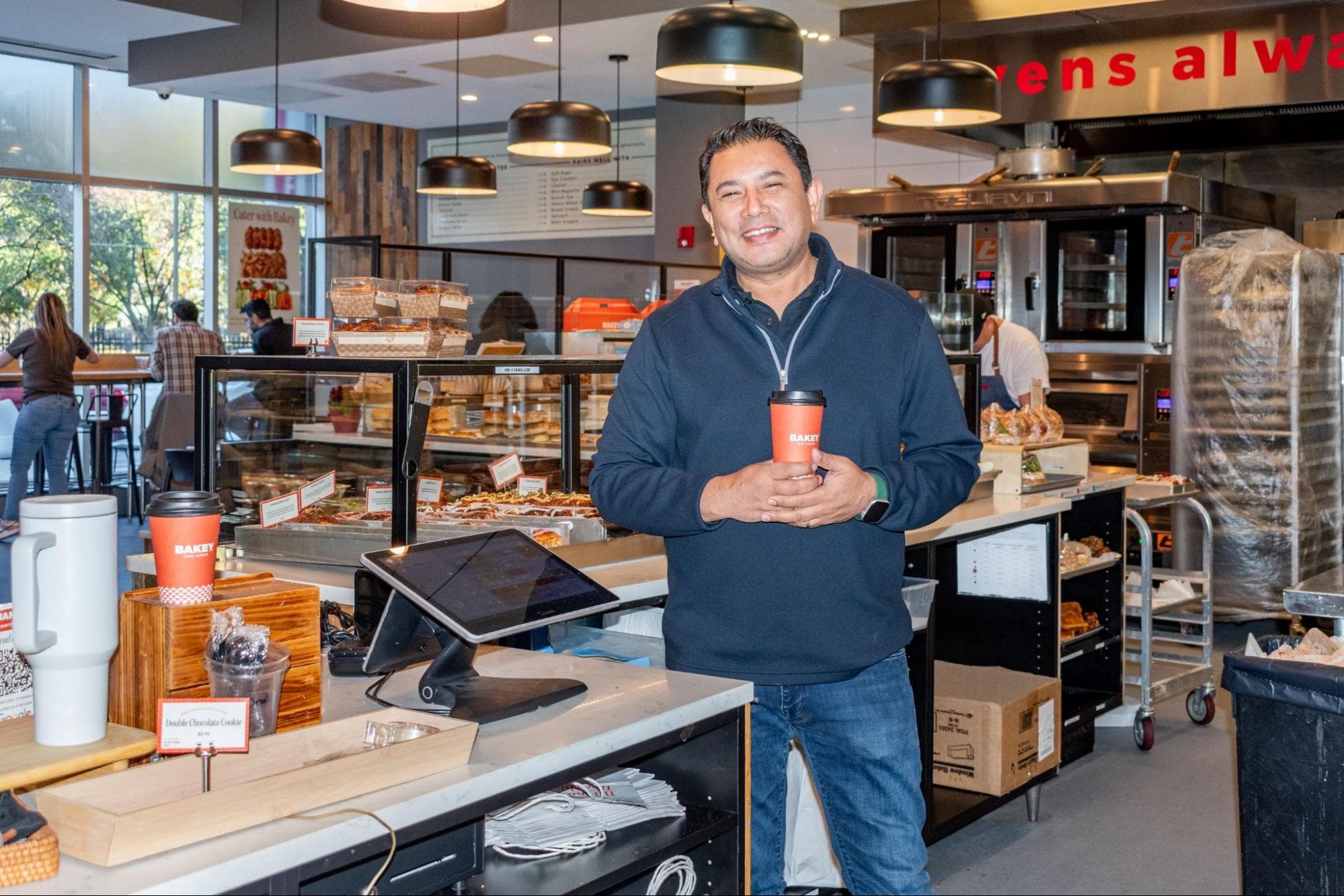 KC Brijesh stands behind the counter at a Bakey shop holding a coffee cup